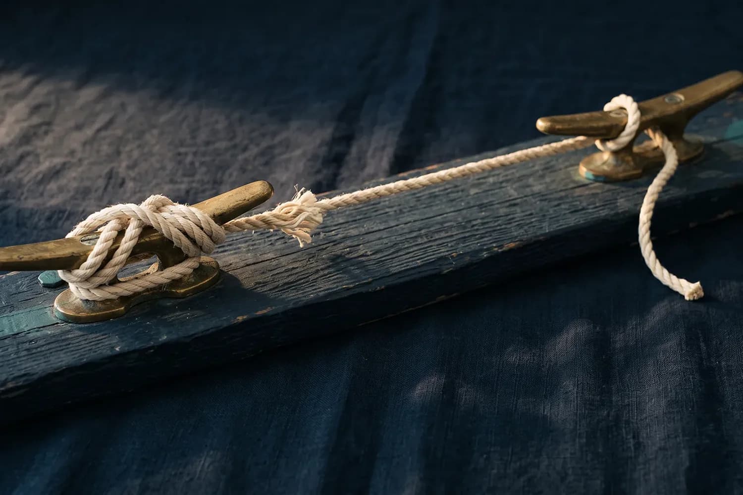 Deux taquets de laiton sur une planche bleue, reliés par un cordage effiloché, serré sur l'un, à peine passé sur l'autre